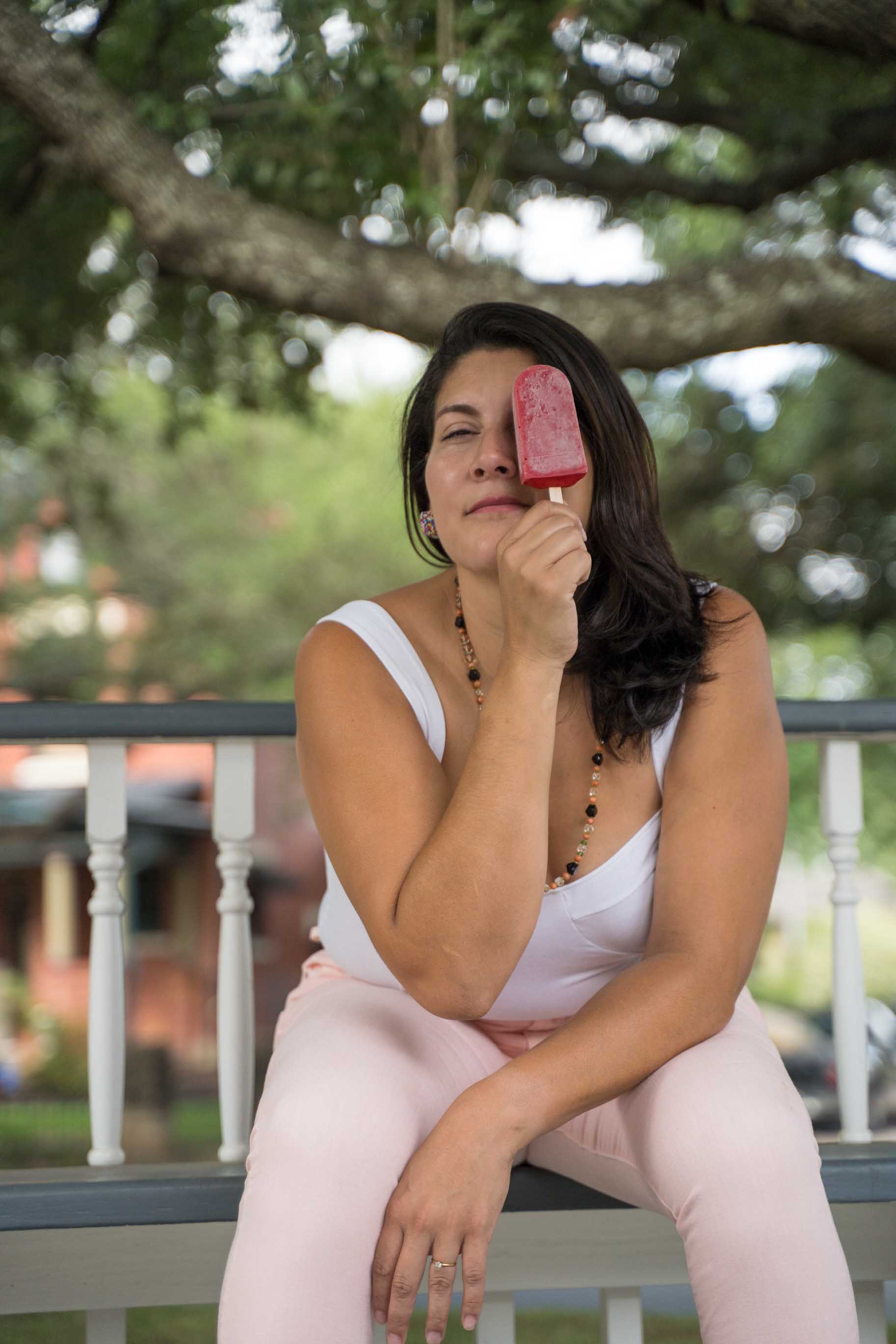 Woman holding a handcrafted popsicle and covering an eye while sitting at a park.